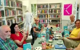 Uczestnicy Dyskusyjnego Klubu Książki, dw&oacute;ch mężczyzn i cztery kobiety. Mają na sobie letnie ubrania z kr&oacute;tkimi rękawami w jasnych kolorach. Siedzą w bibliotece na czarnych, drewnianych krzesłach przy miętowym stole, na kt&oacute;rym są przekąski, napoje, filiża