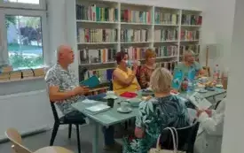 Uczestnicy Dyskusyjnego Klubu Książki, trzy kobiety i trzech mężczyzn. Jeden z nich, ubrany na biało stoi tyłem. Pozostali siedzą przy miętowym stole z przekąskami, napojami, filiżankami, talerzykami. Tr&oacute;jka z nich trzyma książki &bdquo;Ziemianki&rdquo; Marty Strzele