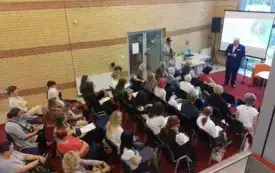 Publiczność podczas Narodowego Czytania. Kilkadziesiąt os&oacute;b siedzi na krzesłach w sali, z przodu, na scenie stoi Dyrektor Biblioteki Sławomir Szenwald, a za nim znajduje się ekran z prezentacją.