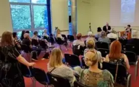 Publiczność zgromadzona w sali, w tle plakat wydarzenia na rzutniku, a przed nim na fotelach siedzi Zofia Mąkosa i dyrektor Sławomir Szenwald