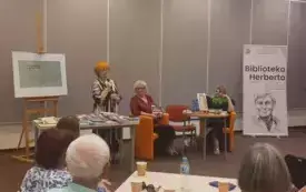 (Od lewej): Barbara Schroeder stoi przed stolikiem z ksiązkami i przemawia do mikrofonu, Marianna Lis i Elżbieta Olszewska-Schilling siedzą przy stole na pomarańczowych fotelach. Przed nimi siedzi publiczność.