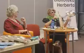 (Od lewej): Marianna Lis i Elżbieta Olszewska-Schilling siedzą przy stole na pomarańczowych fotelach i prowadzą dyskusję. Obok stoi stolik z ksiązkami i baner Biblioteki Herberta.