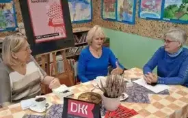 Trzy kobiety siedzą przy stole. Rozmawiają i gestykulują. Na stole jest tabliczka z logo DKK i wrzos w doniczce.