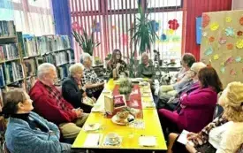 grupa dorosłych os&oacute;b i duży biały pies przy stole, trwa spotkanie DKK, s&oacute;ł ozdobiomy kwiatami, talerz z ciastkami, książki i filiżanki. W tle regały z książkami i duże okna