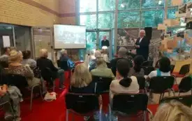 Na pierwszym planie widać kilka rzęd&oacute;w krzeseł, na kt&oacute;rych siedzą osoby odwr&oacute;cone tyłem do fotografa. Przed nimi znajduje się st&oacute;ł z lampką oraz ekran projekcyjny, na kt&oacute;rym jest wyświetlona prezentacja. Przy stole stoją dwie osoby: jedna siedzi, druga st