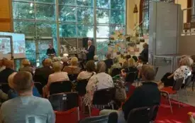 Na zdjęciu widać grupę ludzi siedzących na krzesłach skierowanych w stronę dw&oacute;ch os&oacute;b występujących przy mikrofonach z przodu pomieszczenia. W tle są wysokie okna z widokiem na drzewa,. Pomieszczenie jest jasne, przestronne, z czerwonym dywanem lub wykład