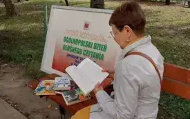 Pani siedzi na ławce i czyta książkę. W tle widoczny park oraz tablica informacyjna.