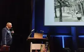 Jeden z panelist&oacute;w przemawiający podczas wykładu. Za nim fragment czarno-białego zdjęcia