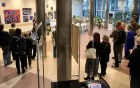 Grupa ludzi stojąca wewnątrz nowoczesnego budynku na wystawie. Widoczne są wysokie okna z dużą ilością światła dziennego oraz obrazy zawieszone na ścianach. Osoby są zwr&oacute;cone plecami do aparatu, większość z nich ubrana jest na ciemno. W tle widać rośliny 
