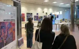 Wnętrze  przestrzeni wystawowej, w kt&oacute;rej kilka os&oacute;b widocznych od tyłu stoi i słucha kobiety, kt&oacute;ra prawdopodobnie otwiera wystawę. Na ścianie za nią znajduje się kilka kolorowych obraz&oacute;w, gł&oacute;wnie w odcieniach niebieskiego, r&oacute;żowego i fioletowego. Po lew