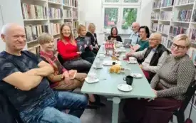 Uczestnicy Dyskusyjnego Klubu Książki siedzą przy miętowym stole. Na stole są słodycze, talerzyki, filiżanki, logo DKK na małej sztaludze. Troje uczestnik&oacute;w spotkania trzyma książki Izabeli Janiszewskiej &bdquo;Noc kłamstw&rdquo; w g&oacute;rze. Po obu stronach za czytel