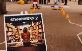 Na pierwszym planie książki oraz plakat z nazwą: &bdquo;Stanowisko 2. Obsługa czytelnik&oacute;w&rdquo;. W tle dzieci pokonujące tor przeszk&oacute;d.
