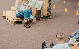 Bibliotekarka czyta książkę przedszkolakom zgromadzonym pod bibliotecznym drzewem.