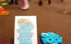 Na pierwszym planie ilustracja kota śpiącego na książkach oraz talerzyk z niebieskimi obrazkami w kształcie książek. W tle biegające dzieci.