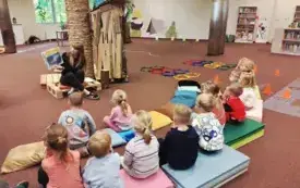 Bibliotekarka czyta książkę przedszkolakom zgromadzonym pod bibliotecznym drzewem.