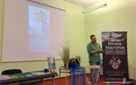 Konrad Hamkało opowiada o swojej książce. Zanim plakat wydarzenia, a przed nim dekoracja - na stole leży dzban, trzcina, a z przodku na podłodze niebieski tiul, na kt&oacute;rym figury syre i książki