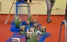 Konrad Hamkało opowiada o swojej książce. Zanim plakat wydarzenia, a przed nim dekoracja - na stole leży dzban, trzcina, a z przodku na podłodze niebieski tiul, na kt&oacute;rym figury syre i książki