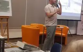 dr Przemysław Pawlak przemawia do mikrofonu, za nim stoi ekran z wyświetlającymi się fotografiami z życia Witkacego.