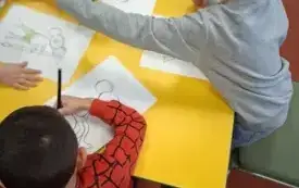 Na stole leżą cztery kartki do kolorowania. Dw&oacute;ch chłopc&oacute;w koloruje wydrukowane &bdquo;Plastusie&rdquo;.