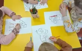 Czworo dzieci siedzi przy stole. Na stole jest kubek z kolorowymi kredkami. Pięciolatki kolorują wydrukowane &bdquo;Plastusie&rdquo;.