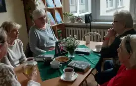 Siedzące przy stole panie, na stole książki, fioletowy emblemat DKK, bukiet jasnych kwiat&oacute;w, filiżanki z napojami, słodycze. W tle brązowa szafa z książkami na niej zielona roślina, obok okno i parapet na nim doniczki z roślinami i czarna torebka.