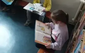 Dzieci siedzące na ławkach z otwartymi książkami w rękach na tle regału z książkami i jasnej ściany z napisem &bdquo;Borejkowo&rdquo;.