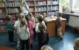 Na tle p&oacute;łek z książkami osoba dorosła trzymająca przed sobą książkę i grupa dzieci. Książki na stole. W tle okno i parapet na kt&oacute;rym zielone rośliny.