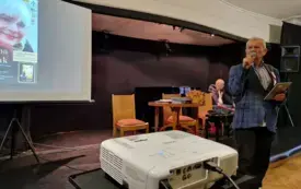 Prezes RSTK &ndash; Czesław Ganda przemawia przez mikrofon. W tle prezentacja multimedialna, wraz z wyświetlanym na ekranie plakatem.