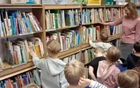 Bibliotekarka oprowadza dzieci po bibliotece. Grupa stoi między regałami z książkami.