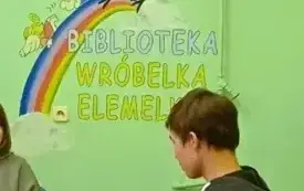 Wnętrze biblioteki, przy stole siedzi klubowicz z książką na kolanach, rozmawia z osobą obok