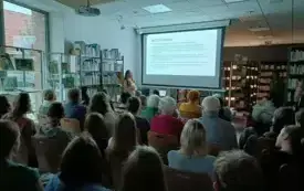 Kobieta stoi przy ekranie z prezentacją wraz uczestnikami siedzącymi na krzesłach.