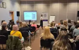 Na zdjęciu widoczna jest sala wypełniona uczestnikami spotkania. Siedzą na krzesłach. W tle widać prowadzącą spotkanie i tablicę, na kt&oacute;rej widać wyświetlony temat spotkania.