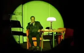 Jerzy Zelnik siedzi przy stole oświetlonym przez lampę. Na scenę rzucana jest okrągła poświata zielonego światła.