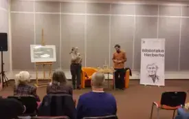 Agnieszka Kopaczyńska-Moskaluk oraz Michał Limboski siedzą na pomarańczowych fotelach i przemawiają do mikrofonu. W tle widać publiczność.