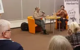 Agnieszka Kopaczyńska-Moskaluk oraz Michał Limboski siedzą na pomarańczowych fotelach i przemawiają do mikrofonu. W tle widać publiczność. 