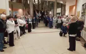 Korytarz biblioteczny, na drugim planie schody prowadzące na piętro. Zdjęcie uczestnik&oacute;w wernisażu, kt&oacute;rzy stoją wzdłuż ścian i na schodach.