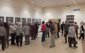 Grupa os&oacute;b na bibliotecznym korytarzu ogląda wiszące na ścianach prace.