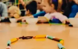 Dzieci przy stolikach tworzą amulety z kolorowego makaronu i koralik&oacute;w.