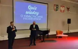 Dyrektor WiMBP Sławomir Szenwald i Patryk Lewandowski prowadzą quiz wiedzy o Paderewskim, w tle fragment prezentacji, fortepian i fotel ze stolikiem kawowym