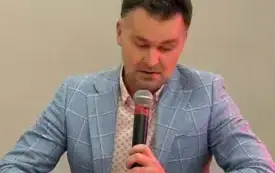 Grzegorz Urbanek siedzi na pomarańczowym fotelu, czyta fragmenty książki, trzymając w jednej ręce książkę, w drugiej mikrofon
