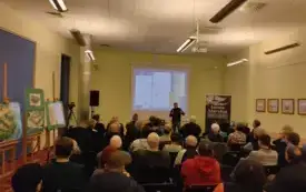 W sali na pierwszym planie zgromadzona publiczność, z lewej strony rysunki Roberta M. Jurgi. Na dalszym planie opowiadający Jurga, wyświetlony slajd oraz wyposażenie sali (glośnik, stolik, rollup Nowej Marchii)