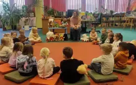 Dzieci siedzą na dywanie w k&oacute;łeczku z bibliotekarką, kt&oacute;ra opowiada o historii powstania pluszowego misia.