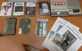 Na jasno brązowym blacie foldery dotyczące wystawy, kasety magnetofonowe i odtwarzacz kaset magnetofonowych.