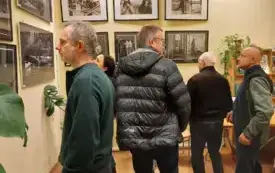 Na jasnej ścianie kolorowe i czarno-białe zdjęcia. W tle regał z książkami i zielone rośliny. Osoby oglądające zdjęcia.