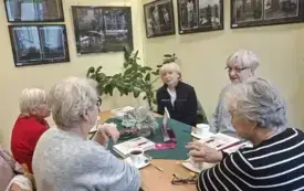 Siedzące przy stole panie, na stole zielona serweta, książki, fioletowy emblemat DKK, rośliny w wazonie, filiżanki z napojami, słodycze, okulary. W tle jasno ż&oacute;łta ściana, na niej wystawa fotografii. W kącie zielona roślina.