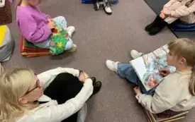 Dzieci wraz z prowadzącą siedzą na dywanie i wsp&oacute;lnie czytają klubową książkę.