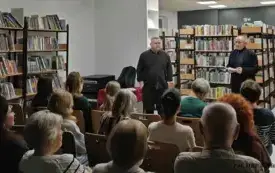 Grupa os&oacute;b siedzi na krzesłach naprzeciwko dw&oacute;ch stojących mężczyzn.