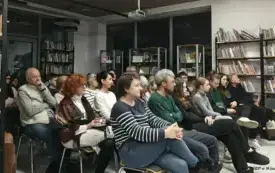 Grupa os&oacute;b siedzi w rzędach na krzesłach w otoczeniu regał&oacute;w z książkami.