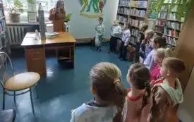 Grupa dzieci z podniesionymi prawymi rączkami i osoba dorosłą trzymająca zieloną kartkę. Przed nią jasno brązowy stolik na nim książki i wielki stojący kartonowy oł&oacute;wek. W tle regały z książkami, jasna ściana z kolorowym symbolem biblioteki, okno i zielon