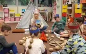 Dzieci siedzą wok&oacute;ł ogniska i słuchają bibliotekarki czytającej książkę.